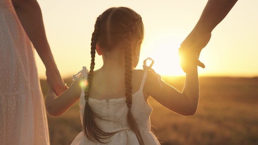 cheerful girl holding her parents' hands at sunrise in a round up of baby girl names that start with...