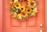 A fall wreath with orange and yellow flowers, golden pumpkins, leaves, and pinecones hanging on an o...