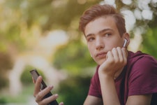 A teenage boy holds his smartphone while looking away into the distance. He does not look super happ...