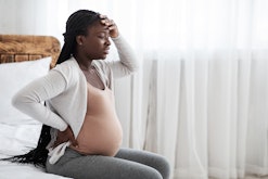 pregnant woman sitting on the edge of her bed with her hand on her back and other hand on her forehe...
