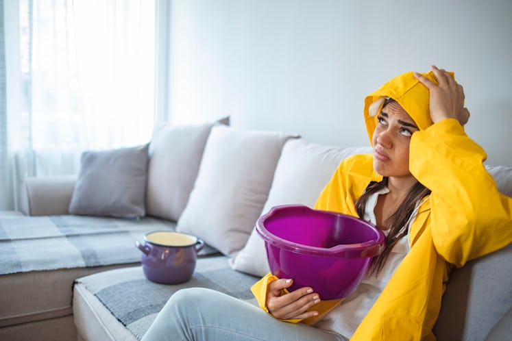 A young woman in raincoat sits on a sofa, holding a bucket to collect dripping water from the ceilin...