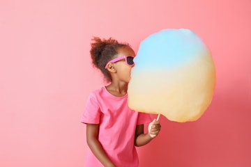 Little African-American girl eating cotton candy on color background