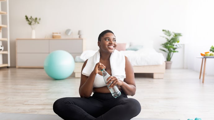 Plump black lady in sportswear sitting on sports mat with bottle of clear water at home, full length...