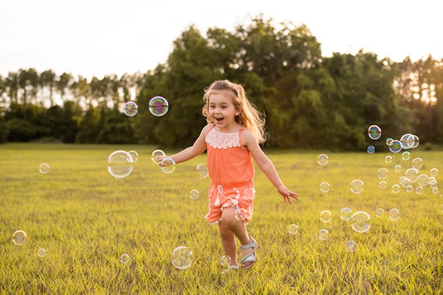 Little girl in orange romper catching soap bubbles on grass in a field at sunset. Xenia is a girls n...
