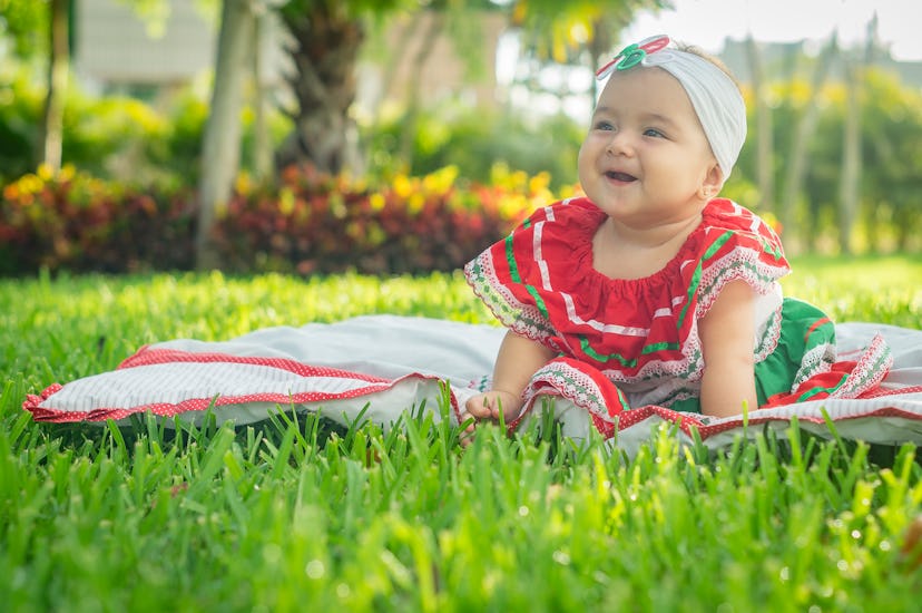 A baby girl sits on a blanket wearing a traditional Mexican dress. The name Xóchitl is a unique girl...