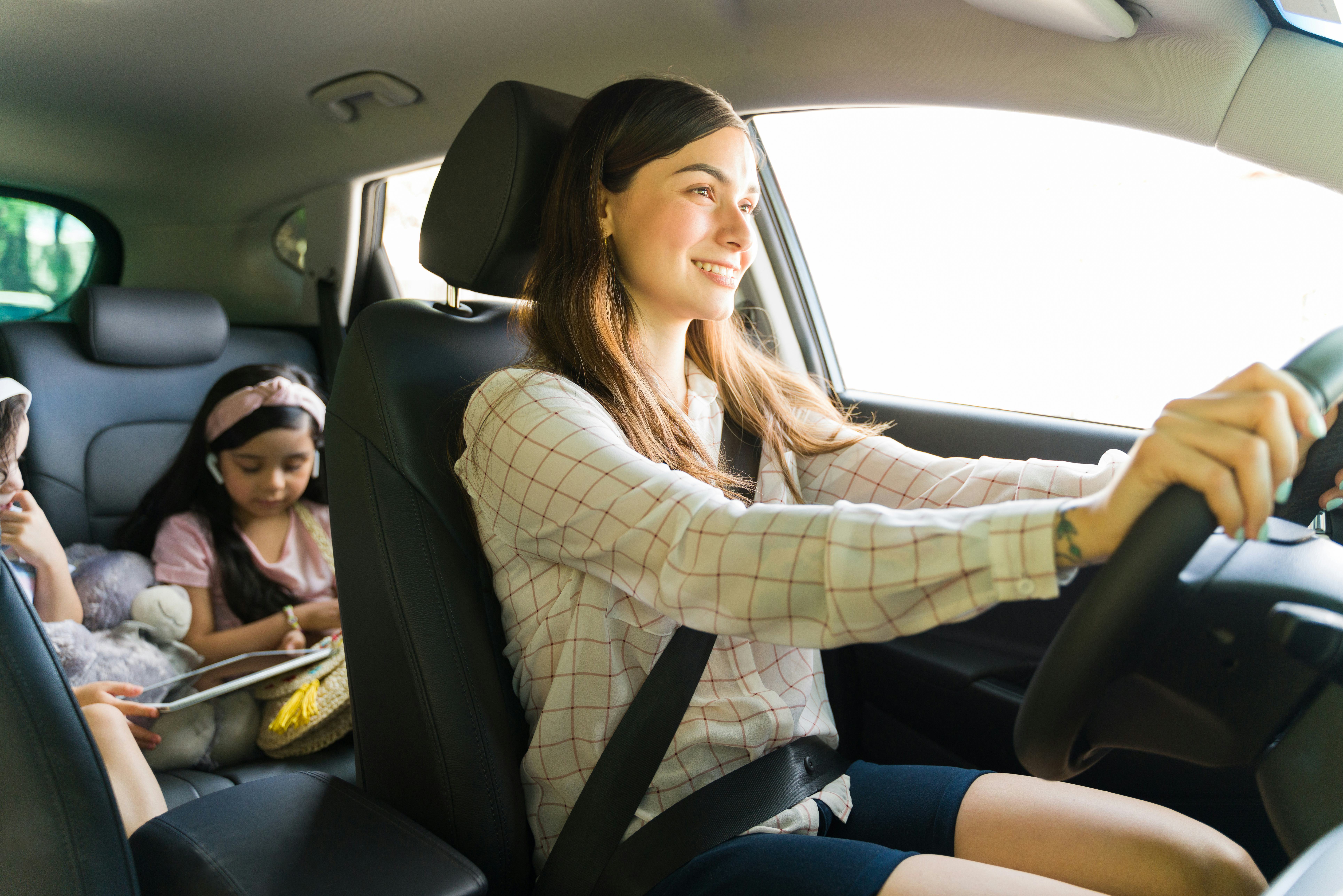 Entertaining my children. Beautiful young mom smiling while driving a car with her kids in the back ...