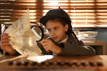 Cute little detective exploring document with magnifying glass at table in office