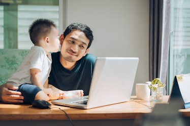 A father works from home at a laptop on his desk while holding his son.