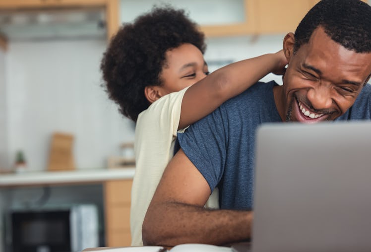 Boy hugging and playing with dad while his dad works from home on a laptop.