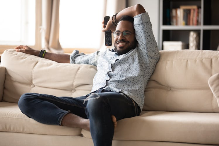 Man relaxing on sofa at home, holding remote controller, watching television in living room.