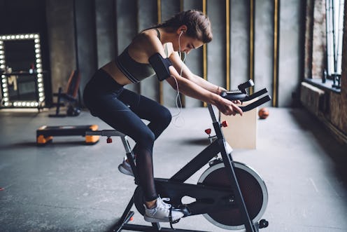 Concentrated fit female in sportswear with dark braided hair burning calories on spin bike and liste...
