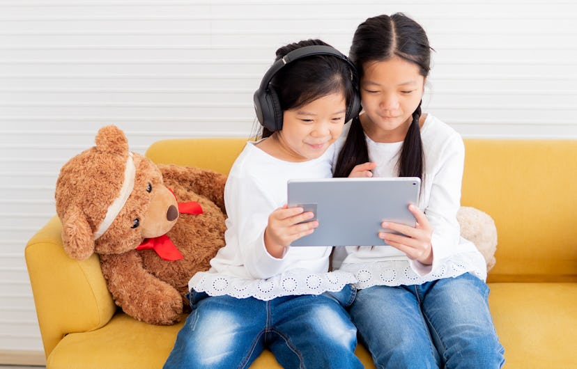 Cheerful two little kid sisters sitting at yellow couch watching online movies or playing games on t...