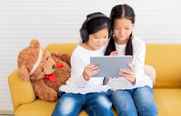 Cheerful two little kid sisters sitting at yellow couch watching online movies or playing games on t...