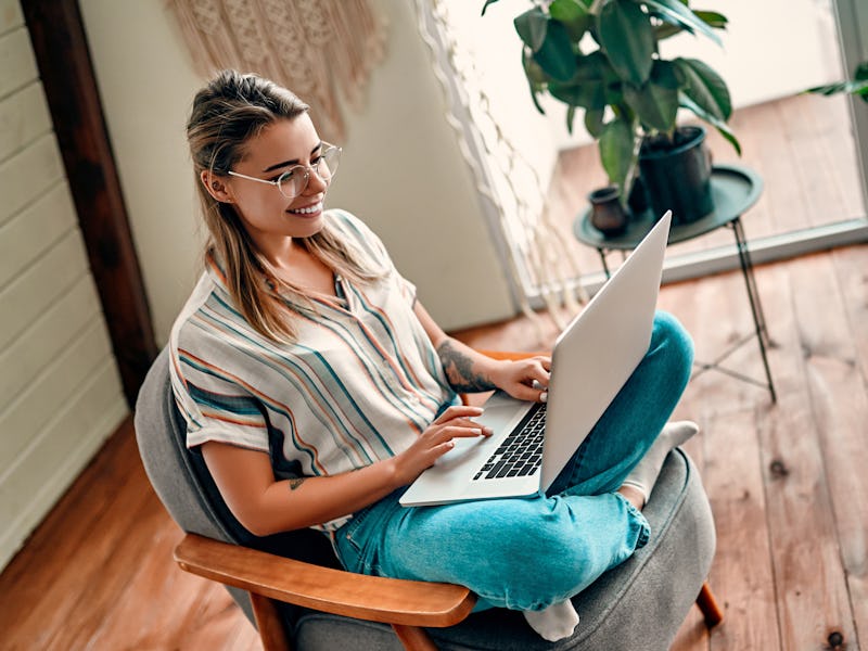 Attractive young woman in glasses works at a laptop while sitting cross-legged in a comfortable chai...