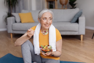Healthy lifestyle concept. Happy senior woman eating fresh vegetable salad, sitting on yoga mat afte...