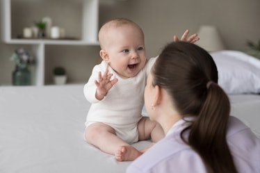 A mom faces her baby who is smiling and sitting on a bed in a white onesie.