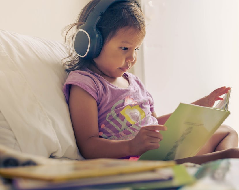 Little girl reading a book at home, wearing headphones. Audio books.