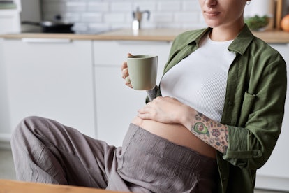 Close up of tattooed pregnant woman relaxing in morning with cup of herbal tea, copy space
