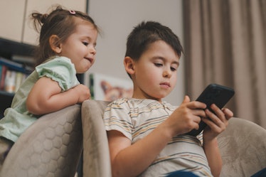 Little adorable brother and sister using smartphone, playing, watching cartoons together. Addicted t...