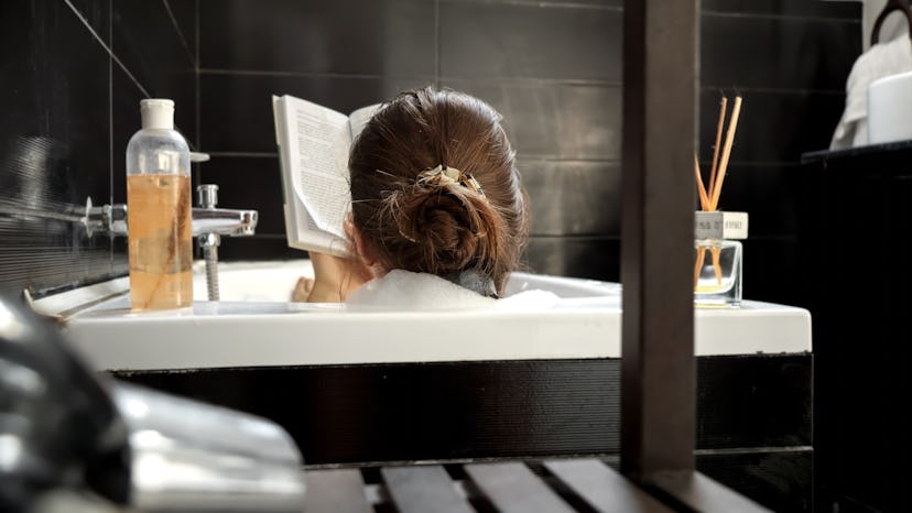 Rear view of elegant woman relaxing in bath with interesting book. Self-care, education, hygiene and...