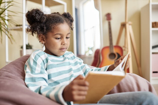 Portrait of little black girl reading book at home sitting in cozy bean bag chair, copy space