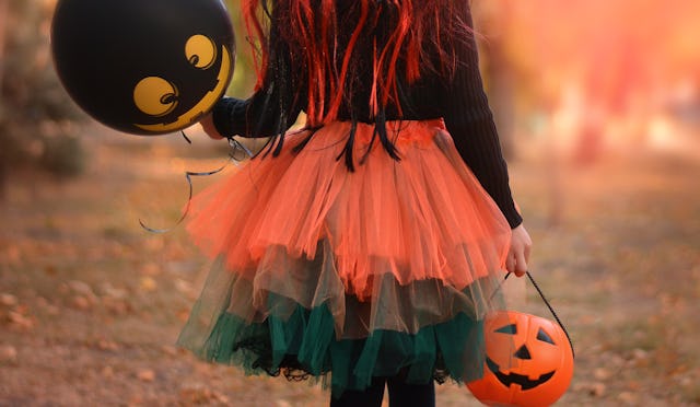 Little girl in witch costume with a balloon and a pumpkin bucket playing in autumn park. Child havin...