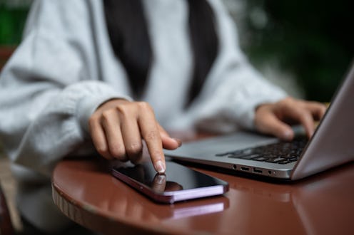 A close-up of a woman tapping on her smartphone screen, checking messages while working on her lapto...