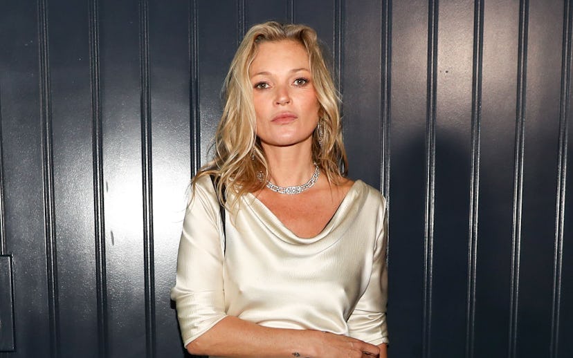 A woman stands against a dark paneled wall wearing a silk cream gown with a black sash and holding a...