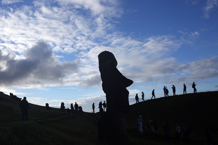 The silhouette of a tall Moai statue's face shows its profile. Behind, a group of people visit the T...