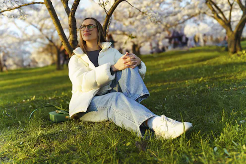 Model Released - Young woman sitting on grass in blooming cherry garden and looking at sakura trees,...