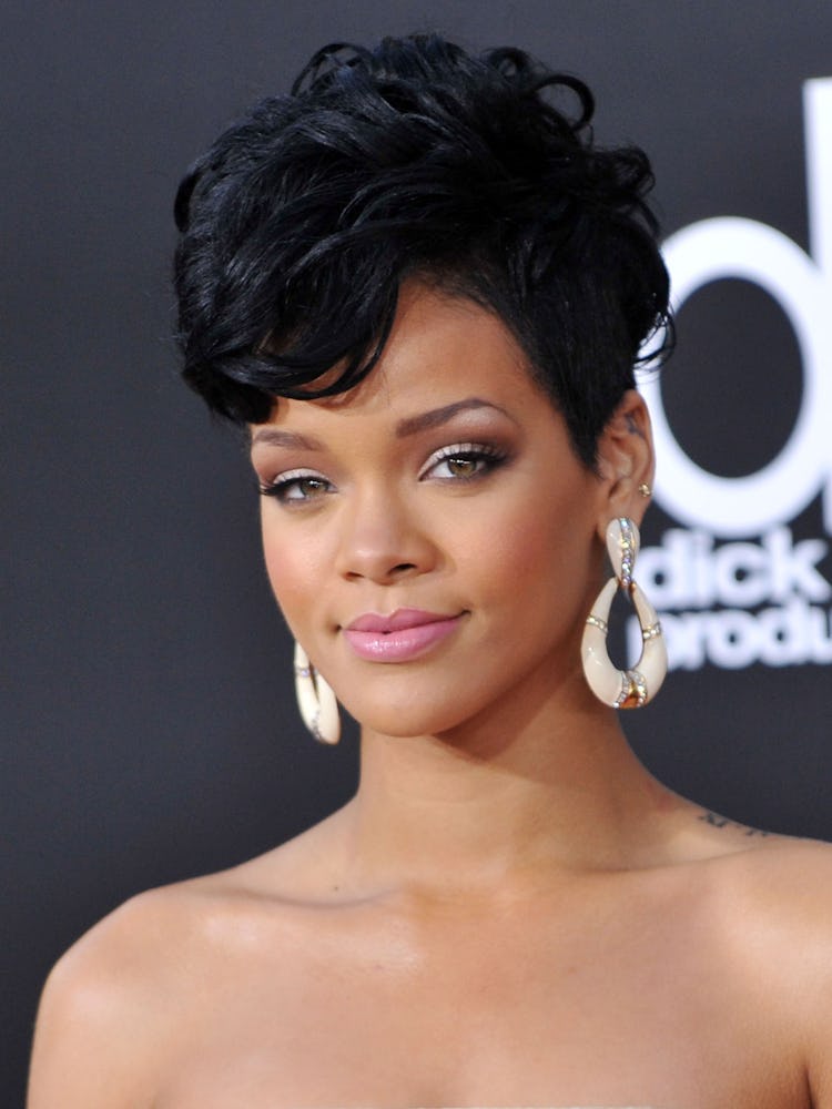 Rihanna with her hair in a jet black curly pompadour, smoky eyes and a nude lip with large white hoo...