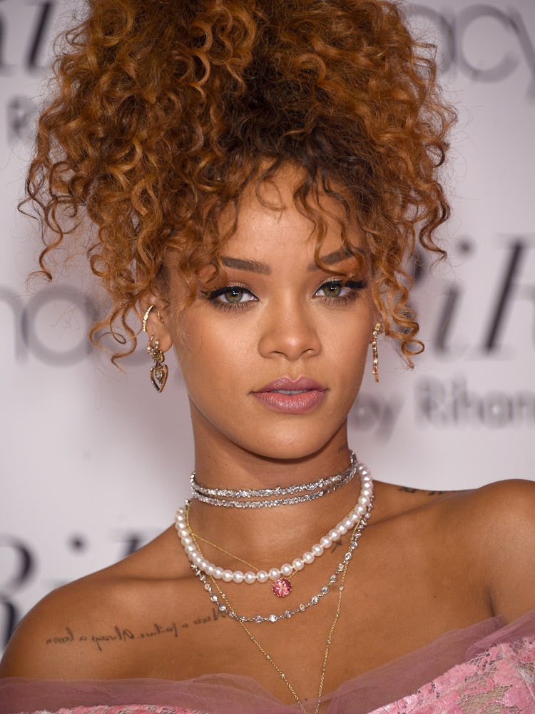 Rihanna with a honey-blonde, curly, high ponytail wearing a pink dress and pearls with a pink gem on...