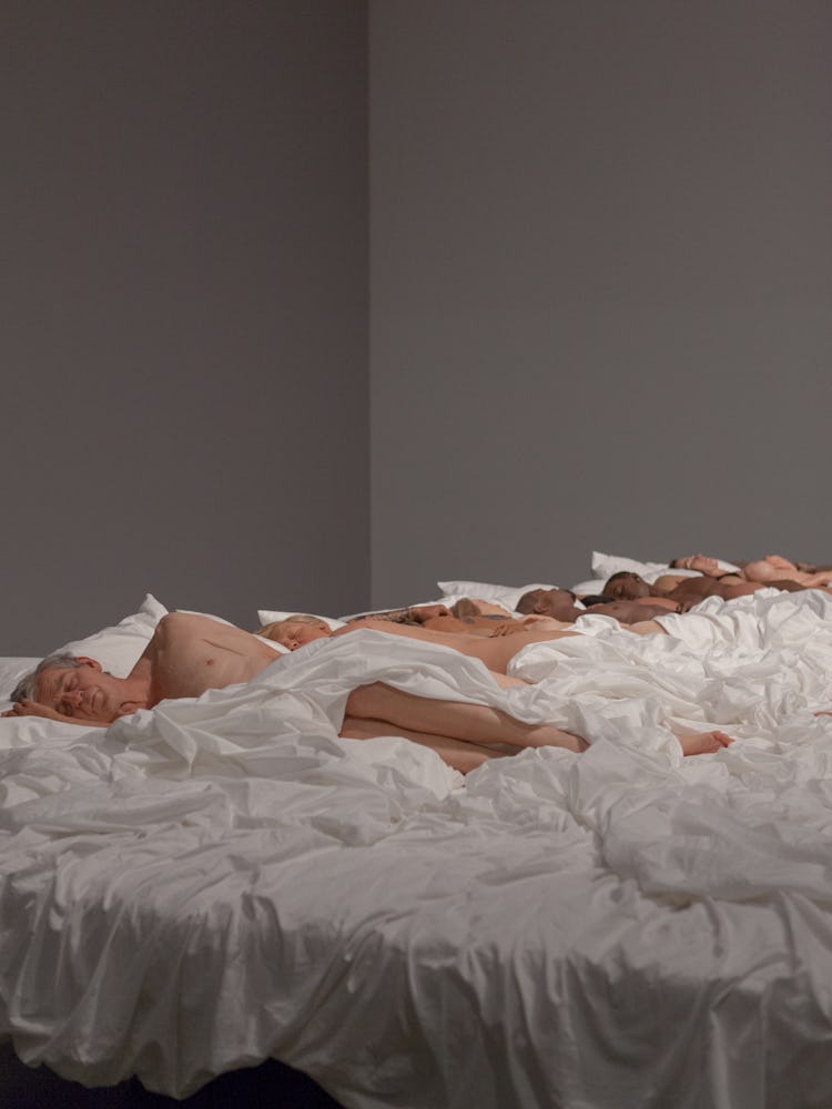 Installation view of Kanye West’s Famous sculpture with doll bodies lying in a bed with white sheets