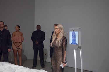 Kim Kardashian standing next to a small tv monitor with Kanye West visible on it in the gallery