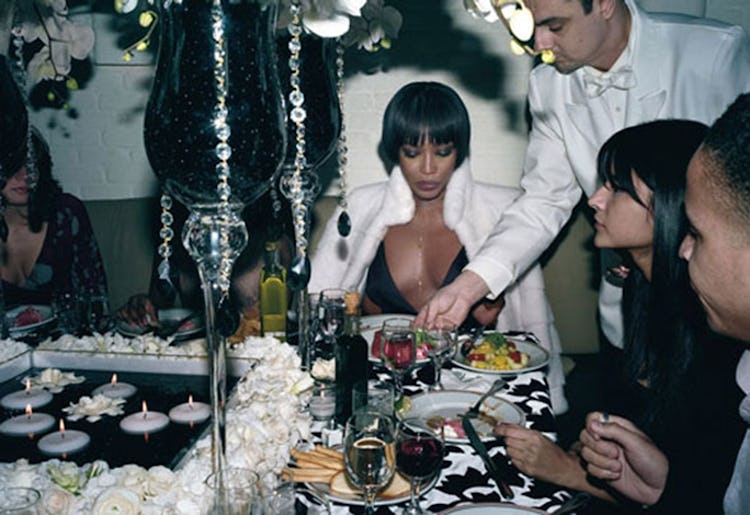 Naomi Campbell sitting in a black dress and white jacket and eating food from a dinner table
