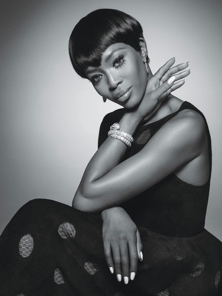 Naomi Campbell with short hair sitting and posing in a black dress and wearing diamond jewelry