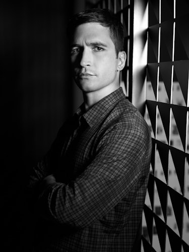 A man in a dark room with his arms crossed while wearing a checked shirt