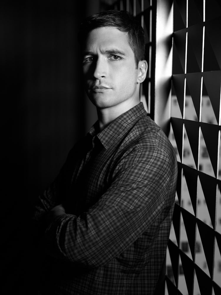 A man in a dark room with his arms crossed while wearing a checked shirt