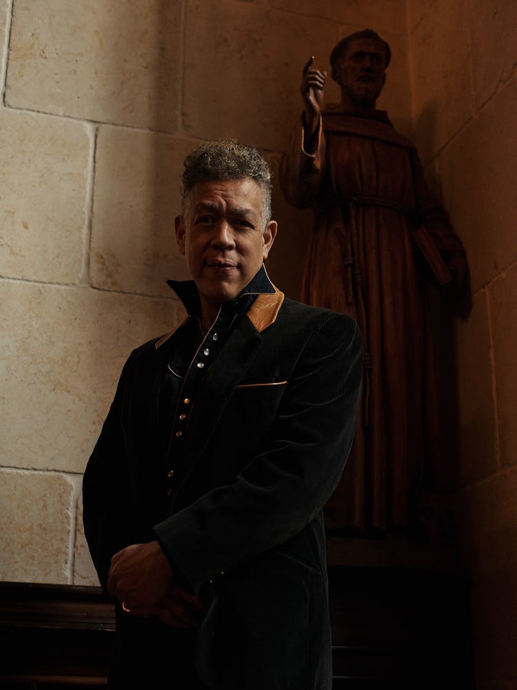 A man standing in a black jacket with a yellow collar and a sculpture behind him