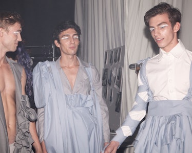 Male models in light blue and grey dresses, with bright white eyeshadow waiting backstage at VFiles'...