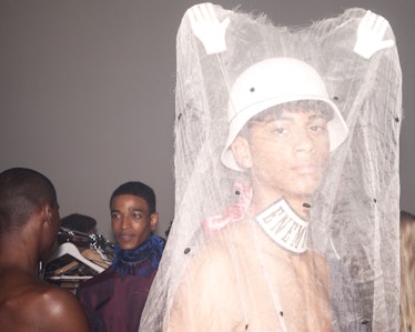 A model backstage at the show wearing a hat with hands on top of it and lace draped over the hands
