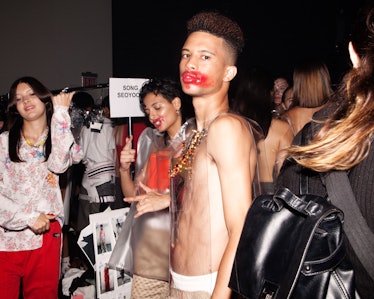 A male model with red lipstick smudged all over his lips and cheeks standing among other models back...