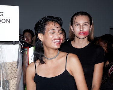 Two models in black tops with red lipstick smudged on their faces backstage at the show 