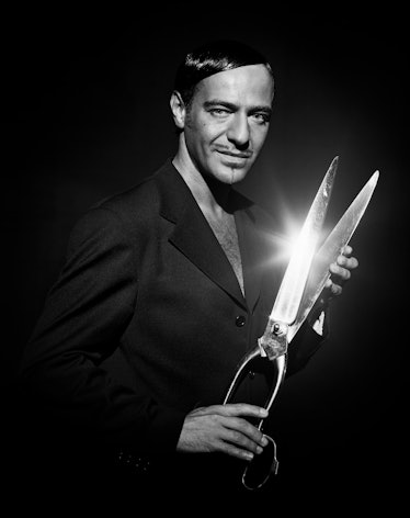 John Galliano posing for a photo in a black blazer while holding giant scissors 