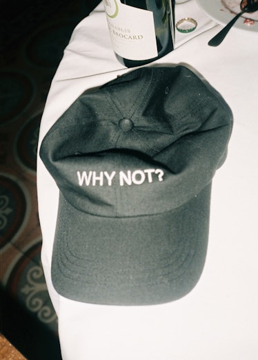 A black cap that says "Why Not?" on it, placed on a white table next to a bottle of wine in Vetemant...