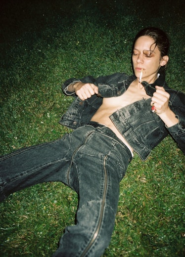 A model lying in the grass smoking a cigarette and wearing a denim jacket and matching jeans 