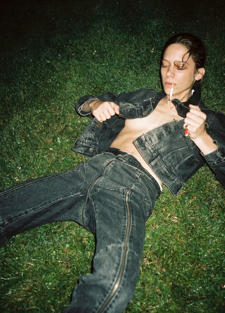 A model lying in the grass smoking a cigarette and wearing a denim jacket and matching jeans 