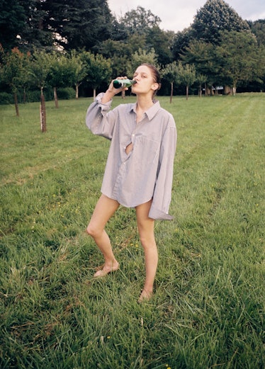 A model in Vetements Summer Camp standing in a field in an oversized button-up, drinking a beer 