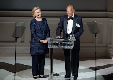 Hillary Clinton in a navy blazer and trousers standing next to a man who is giving a speech in June ...