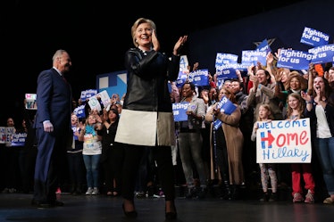 Hillary Clinton in a black-white coat and black trousers clapping and walking in front of  a crowd i...